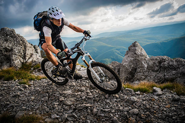 Mountain biker riding his bike down a rocky pathway with sharp grey rocks and ominous black clouds overshadowing green wavy hills in the background.