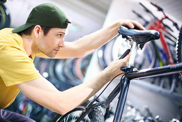 Early 30's bike mechanic tightening seat on customer's bike. Wearing yellow shirt and backwards pointed baseball cap.