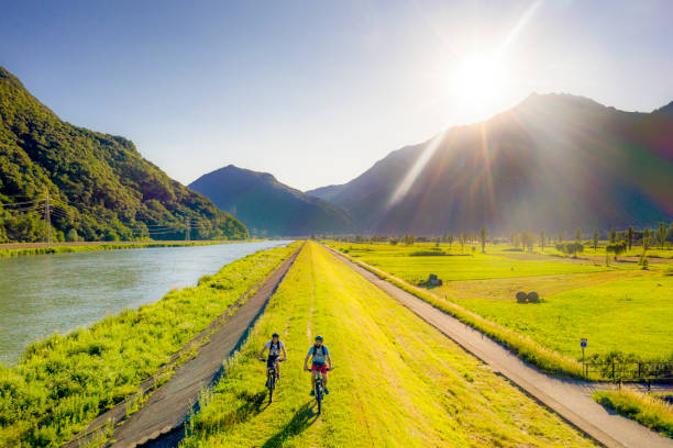 Two cyclist ride e-bike on the riverside of Adda river. Valtellina, Italian Alps