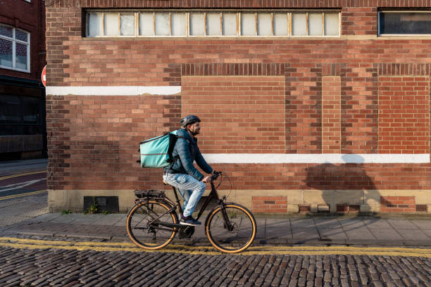 A delivery man wearing a bicycle helmet and food delivery bag, riding his bike on a cobbled road in Newcastle upon Tyne, England. He is on his way to deliver food to a customer during dusk.