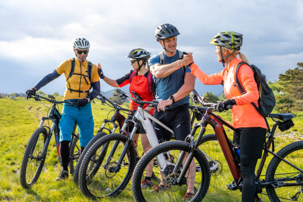 Mature couples shaking hands and gesturing while standing with electric bikes on mountain top against cloudy sky.