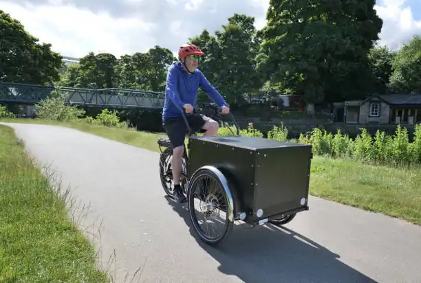 Middle aged white man cycles on a large black electric cargo bike on a tarmac lane through a lush green park