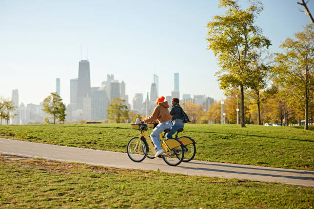 Full length view of vacationers in 20s and 30s, dressed warmly and pedaling near Lake Michigan shore, urban skyline in background.